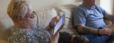 Middle-aged couple sat on a sofa, one reading an ipad with a dog on their lap.